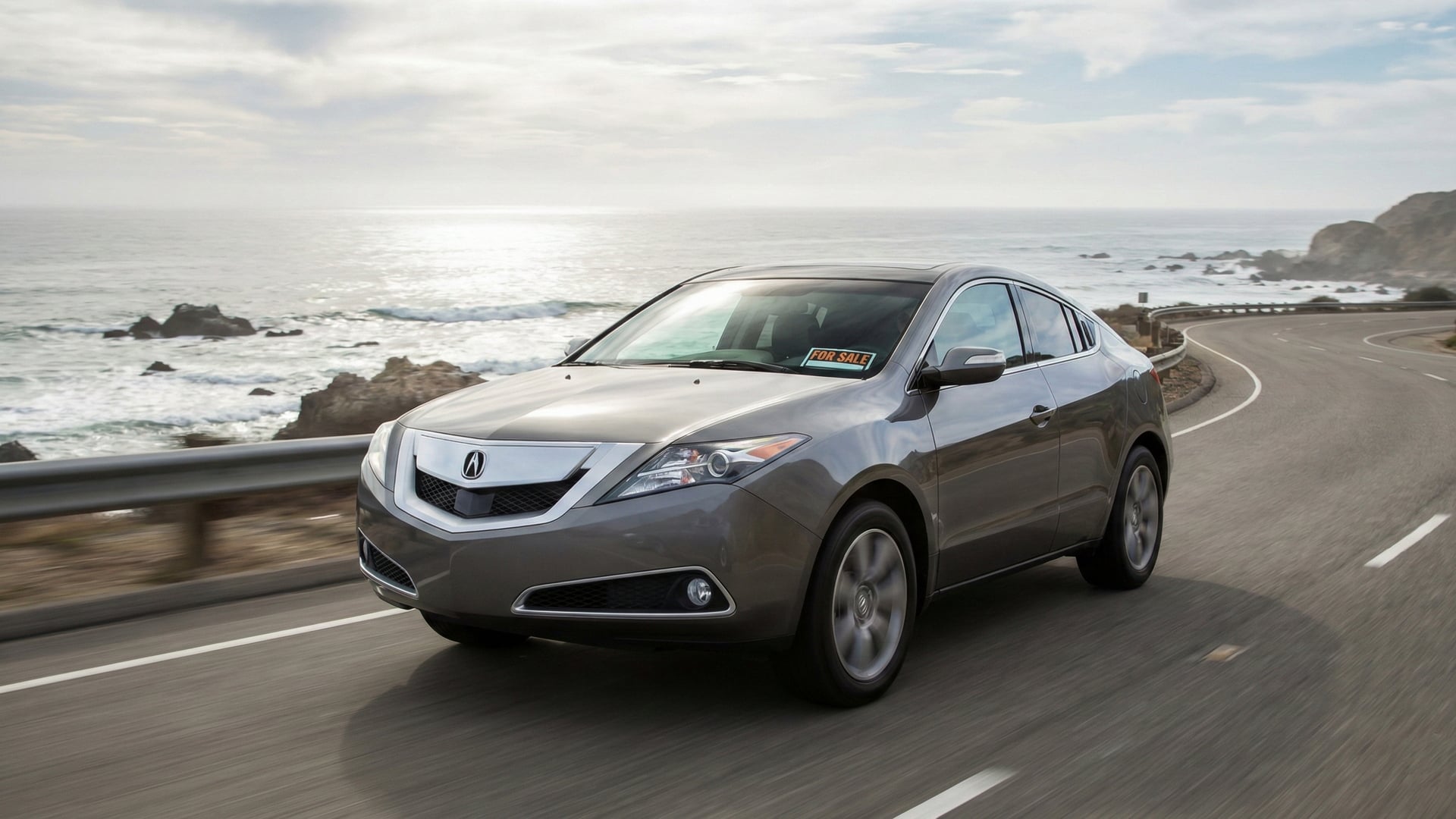 Acura ZDX in gray driving along a California coastal highway with a for-sale sign in the windshield.
