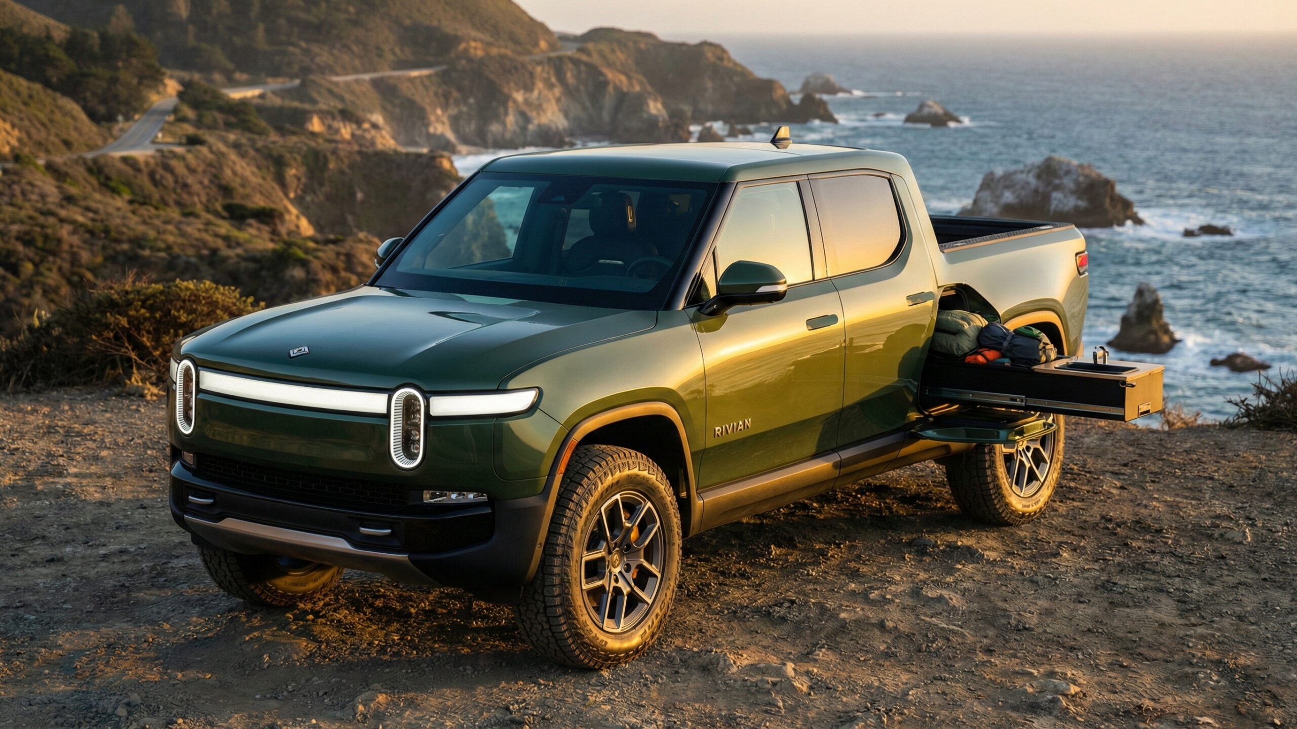 Green Rivian pickup truck with gear tunnel drawer open on a California coastal cliff at sunset.
