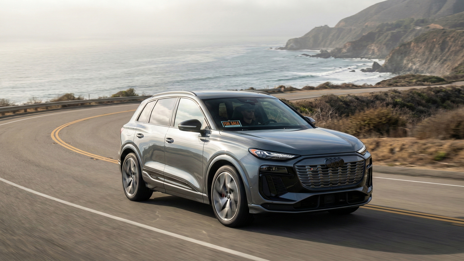 Audi SQ6 e-tron silver SUV driving along a California coastal highway with a For Sale sign in the windshield.