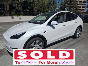 Front three-quarter view of a white Tesla Model Y electric crossover utility vehicle parked on a concrete lot with a promotional banner overlay, showing how to sell my Tesla Model Y for more to CarZilo.